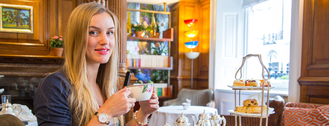 Eine junge Frau sitz in einem englischen Café und trinkt Tee, auf einer Etagere leigen Kuchen und Scones