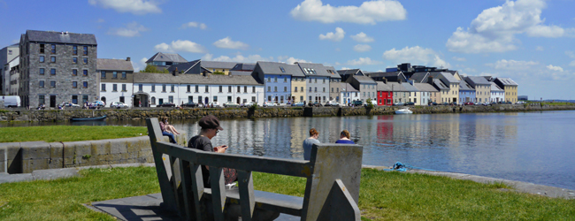 Menschen sitzen an der Promenade und entspannen im Sonnenschein vor den Häusern von Galway