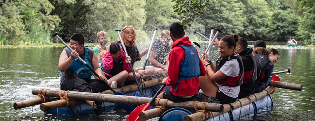 Schüler sitzen in einer Art Boot aus Fässern und paddeln über einen See bei Adventure England